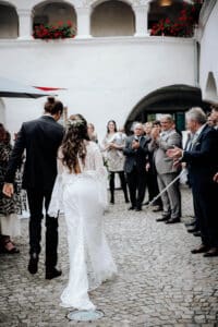 Braut und Bräutigam gehen Hand in Hand einen gepflasterten Weg entlang, umgeben von applaudierenden Gästen auf beiden Seiten. Die Braut trägt ein langes weißes Spitzenkleid mit weiten Ärmeln. Diese malerische Hochzeitsszene wird von weißen Bögen und roten Blumen eingerahmt, die Gäste sind in Abendgarderobe gekleidet.