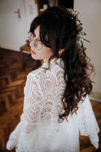 Eine Frau mit langen, lockigen Haaren, die mit kleinen Blumen geschmückt sind, trägt eine Brille und ein zartes Spitzenkleid und blickt nach oben in die Kamera. Sie steht auf einem Holzboden, der an eine romantische Hochzeitskulisse erinnert, mit einer Tür im Hintergrund.