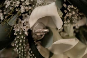 Nahaufnahme eines Paars Eheringe, eingebettet zwischen zarten weißen Blumen und üppigen grünen Blättern, die die Essenz der Hochzeitsplanung einfangen.