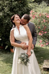 Ein lächelndes Paar umarmt sich an seinem Hochzeitstag in einem Garten. Die Frau in einem ärmellosen weißen Kleid hält einen Blumenstrauß, während sich der Mann im grauen Anzug liebevoll zu ihnen beugt. Rosa Blumen und Grün umgeben sie in diesem sonnigen Moment ihrer Hochzeitsplanung.