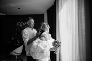 Ein Schwarzweißfoto eines glücklichen Paares an seinem Hochzeitstag an einem Fenster. Die Frau, die einen Blumenstrauß hält, lehnt sich lachend zurück, während der Mann lächelnd hinter ihr steht. Sanftes Licht von draußen beleuchtet sie und verstärkt ihre freudigen Ausdrücke, während sie diesen Moment der Hochzeit genießen.