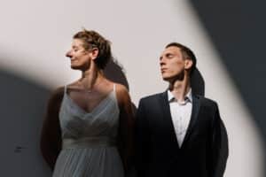 Eine Frau in einem weißen Kleid und ein Mann in einem dunklen Anzug stehen Schulter an Schulter vor einer weißen Wand und denken vielleicht über ihre Hochzeit nach. Sie blicken in entgegengesetzte Richtungen, dramatische Schatten verleihen diesem entscheidenden Moment ihrer Heiratsreise eine geheimnisvolle Aura.
