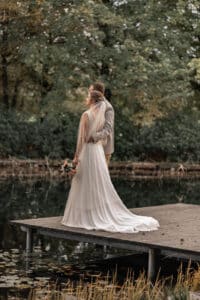 Ein Paar steht auf einem Steg an einem See, umgeben von üppigen Bäumen. Die Frau im weißen Brautkleid hält einen Blumenstrauß, während der Mann im Anzug sie von hinten umarmt. Ihre romantische Szene am ruhigen Wasser verkörpert ihre Traumhochzeit und markiert den Beginn ihres neuen gemeinsamen Lebens.