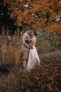 Ein Paar umarmt sich auf einem Feld, umgeben von Herbstlaub, und fängt die Essenz einer perfekten Hochzeit ein. Die Frau im weißen Kleid hält einen Blumenstrauß, während der Mann eine graue Jacke und braune Hosen trägt. Sie stehen eng beieinander unter orangefarbenen Blättern und schaffen eine bezaubernde Atmosphäre für ihre Hochzeitsreise.