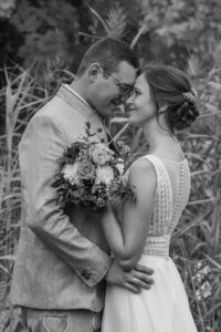 Ein Schwarzweißfoto zeigt eine Hochzeitsszene, in der Braut und Bräutigam Stirn an Stirn stehen. Sie hält einen Blumenstrauß und trägt ein Spitzenkleid, während er einen Anzug trägt. Umgeben von hohem Gras, das die intime, natürliche Atmosphäre ihres besonderen Tages unterstreicht.