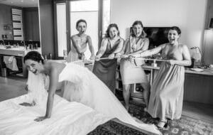 Eine Braut im Hochzeitskleid lehnt sich verspielt auf ein Bett, während fünf Brautjungfern in einem Hotelzimmer enthusiastisch an der Schleppe ihres Kleides ziehen. Alle lächeln und lachen und schaffen so eine lebhafte und fröhliche Hochzeitsszene. Das Bild ist in Schwarzweiß.