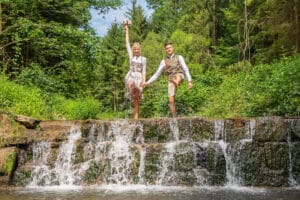 Ein Paar in traditioneller Kleidung posiert fröhlich an einem kleinen Wasserfall in einem üppigen Wald und verkörpert den perfekten Moment für Hochzeitserinnerungen. Die Frau hält Blumen hoch, während der Mann neben ihr steht. Beide zeigen inmitten des Grüns und des fließenden Wassers glückliche Gesichter.