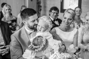 Ein Schwarzweißfoto fängt die Freude einer Hochzeit ein. Ein Mann im Anzug hält ein kleines Kind, das mit einem Blumenkranz geschmückt ist; ein weiteres Kind beugt sich vor. Neben ihnen lächelt eine Frau in einem weißen Kleid, umgeben von elegant gekleideten Gästen, die an der Feier teilnehmen.