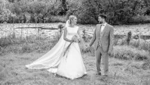 Braut und Bräutigam lächeln sich Hand in Hand neben einem ruhigen Teich in einer üppigen Umgebung im Freien fröhlich an. Ihr elegantes Kleid und ihr Schleier bilden einen Kontrast zu seinem hellen Anzug und verkörpern zeitlose Romantik. Dieser schwarz-weiße Hochzeitsmoment wird wunderschön von den umliegenden Bäumen und dem Gras eingerahmt.