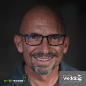 Ein kahlköpfiger Mann mit Brille, Spitzbart und warmem Lächeln, der eine dunkle Jacke trägt. In den unteren Ecken befinden sich zwei Logos: „perfektheiraten harald winkler“ links und „Austrian Wedding Professional“ rechts, die professionelle Hochzeitsplanung für Ihre Traumhochzeit verkörpern.
