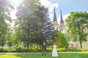 Ein Paar steht Hand in Hand auf einer Wiese und bereitet sich auf seine Hochzeit vor. Im Hintergrund ragt eine Kirche mit zwei Türmen vor einem klaren blauen Himmel empor. Sonnenlicht fällt durch die Blätter und erzeugt im Vordergrund Lichtreflexe.