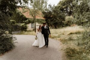 Braut und Bräutigam gehen Arm in Arm auf einem geschwungenen Weg, umgeben von Grün, und verkörpern damit die Essenz einer perfekten Hochzeit. Die Braut trägt ein weißes Kleid und hält einen Blumenstrauß, während der Bräutigam einen dunklen Anzug trägt. Ein rustikales Gebäude und ein Eisentor vervollständigen diese idyllische Kulisse für ihren besonderen Tag.