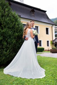 Braut und Bräutigam stehen draußen auf einer Wiese. Die Braut trägt ein weißes Kleid und hält einen Blumenstrauß, während der Bräutigam einen dunklen Anzug trägt. Sie posieren vor einem gelben Gebäude mit schrägem Dach und halten den Moment ihrer Traumhochzeit fest. Links ist ein großer Busch.