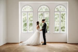 Braut und Bräutigam stehen sich vor drei großen Bogenfenstern gegenüber, draußen ist viel Grün. Die Braut trägt ein langes weißes Kleid und der Bräutigam einen Anzug, was die Eleganz ihrer Hochzeit verkörpert. Sie befinden sich in einem hellen, minimalistischen Raum mit Hartholzboden.