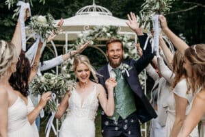 Ein fröhliches Paar feiert nach seiner Hochzeit. Umgeben von Freunden und Familie mit Blumensträußen gehen sie Hand in Hand unter einer Laube hindurch. Der Bräutigam trägt eine grüne Weste und einen Anzug, die Braut ein weißes Spitzenkleid. Alle lächeln.