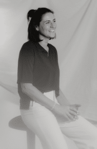 Schwarz-Weiß-Foto einer Frau, die auf einem Hocker vor einem weichen, drapierten Hintergrund sitzt und an eine elegante Braut inmitten der Hochzeitsplanung erinnert. Sie lächelt, trägt ein dunkles kurzärmliges Oberteil und helle Hosen, ihre Hände ruhen auf ihrem Schoß. Ihr Haar ist teilweise hochgebunden.