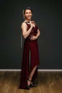 Eine Frau in einem roten ärmellosen Kleid steht auf einem Holzboden vor einem dunklen Hintergrund und erinnert an die Eleganz einer Hochzeit. Sie hält einen Säbel, dessen Spitze auf dem Boden ruht und dessen Schaft anmutig an ihrer Schulter anliegt, als ob sie ihren nächsten Schritt in der großen Hochzeitsplanung des Lebens plant.