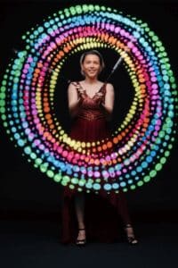 Eine Person in einem roten Kleid führt bei einer Hochzeit eine faszinierende Lichtshow durch, bei der sie mit LED-Poi bunte, kreisförmige Muster vor dem dunklen Hintergrund erzeugt und so dem Ereignis Magie verleiht.