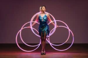 Eine Frau in einem blauen Kleid begeistert die Gäste mit ihrer Darbietung, bei der sechs leuchtende Hula-Hoop-Reifen symmetrisch um sie herumwirbeln. Auf einem Holzboden vor einem sanft beleuchteten rosa Hintergrund verleiht diese magische Darbietung der Atmosphäre jeder Hochzeitsfeier bezaubernde Eleganz.
