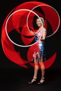 Eine Person in einem Paillettenkleid dreht anmutig zwei leuchtend rote Reifen vor einem dunklen Hintergrund und erzeugt so lebendige kreisförmige Muster, die an die energiegeladene Stimmung einer Hochzeitsfeier erinnern.