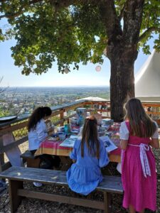 Drei Mädchen sitzen an einem Picknicktisch unter einem großen Baum und sind in Aktivitäten vertieft. Der Tisch ist mit Bastelutensilien für kreative Hochzeitsplanung bedeckt. Im Hintergrund erstreckt sich eine malerische Landschaft unter einem klaren blauen Himmel.