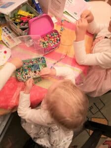 Drei Kinder sitzen an einem Tisch voller bunter Perlen und träumen vielleicht von einem zukünftigen Heiratsantrag. Sie fädeln Perlen auf Schnüre und basteln konzentriert Armbänder oder Halsketten. Der Tisch ist mit einer gemusterten Tischdecke bedeckt und verschiedene Bastelutensilien liegen verstreut herum.