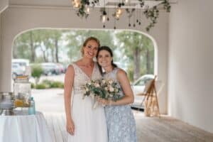 Zwei Frauen stehen lächelnd in einer halboffenen Umgebung zusammen und feiern eine Hochzeit. Eine trägt ein weißes Kleid und hält einen Blumenstrauß, während die andere ein hellblaues gemustertes Kleid trägt. Hängende Lichter und Grünpflanzen schmücken wunderschön die Decke.
