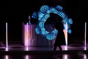Silhouette einer Person vor einem farbenfrohen Lichtspiel mit kreisförmigen blauen Mustern. Sie steht auf einer Bühne mit neonbeleuchteten Säulen und schafft eine dynamische und futuristische Atmosphäre, ähnlich der Aufregung bei der Planung einer Hochzeit inmitten lebhafter Feierlichkeiten.