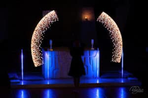 Eine Person steht vor einem Altar mit blauem Licht und beschwört die Magie einer Traumhochzeit herauf. Zwei gebogene, beleuchtete LED-Panels zeigen ein feuerwerksähnliches Muster und erzeugen einen künstlerischen und lebendigen Hintergrund, der zur bezaubernden Atmosphäre dieses Meisterwerks der Hochzeitsplanung beiträgt.