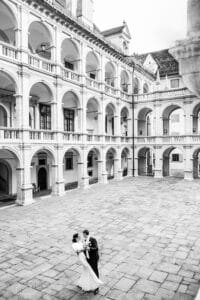 Ein Paar im Hochzeitskleid tanzt fröhlich im Innenhof eines historischen, mehrstöckigen Gebäudes mit großen Bögen und Säulen. Die in Schwarzweiß festgehaltene Szene hebt die Architektur und ihr Hochzeitsglück wunderschön hervor.