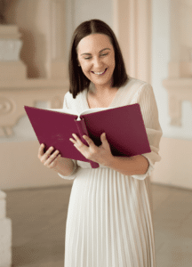 Eine Frau in einem weißen Kleid lächelt, während sie ein magentafarbenes Buch liest. Sie steht drinnen vor einem hellen, eleganten Hintergrund und erinnert an die Anmut einer Hochzeitsplanung. Ihr Haar ist schulterlang und glatt.