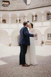 Ein Paar in formeller Hochzeitskleidung umarmt und küsst sich in einem sonnigen, offenen Innenhof mit weißen Bögen und Steinsäulen. Das lange weiße Kleid der Braut und der marineblaue Anzug des Bräutigams schaffen eine zeitlose Liebesszene. Blumen in Töpfen sorgen für einen Hauch von Farbe, perfekt für jede Hochzeitsplanung.