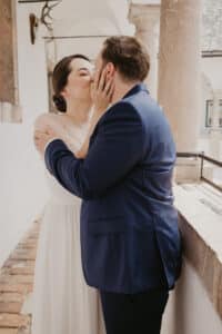 Ein Paar in Hochzeitskleidung küsst sich auf einem Steinbalkon und verkörpert damit die Essenz ihrer Hochzeit. Die Frau trägt ein weißes Kleid und der Mann einen blauen Anzug. Sonnenlicht erhellt die Szene und die Geweihe verleihen diesem bezaubernden Moment eine rustikale Note.
