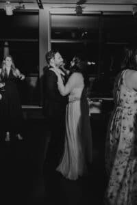 Ein Schwarzweißfoto zeigt ein Paar, das bei einer schwach beleuchteten Hochzeit in einem Innenbereich eng umschlungen tanzt. Der Mann trägt einen Anzug und die Frau ein langes Kleid. Im Hintergrund sind weitere Gäste zu sehen, von denen einige für die Feier ihrer Hochzeit gekleidet sind.