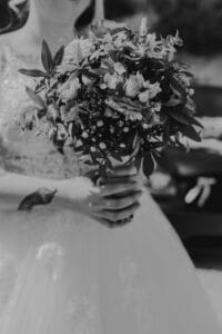 Eine Braut im Hochzeitskleid hält einen großen Blumenstrauß und fängt damit die Essenz einer Hochzeit ein. Das Schwarzweißbild betont die komplizierten Details des Kleides und der Blumen und bietet zeitlose Inspiration für diejenigen, die sich intensiv mit der Hochzeitsplanung beschäftigen.