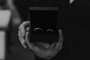 Eine Person, die an der Hochzeitsplanung beteiligt ist, hält eine offene Ringschachtel mit zwei Ringen in der Hand. Das Schwarz-Weiß-Bild betont den Kontrast zwischen ihren tätowierten Händen und den Ringen und fängt einen zeitlosen Moment auf dem Weg zur Hochzeit ein.