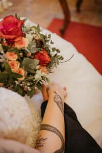 Nahaufnahme der Hände einer Person mit einem Schlangentattoo, die auf einem weißen Kleid ruhen und einen leuchtenden Strauß roter und rosa Rosen halten – perfekt für jede Hochzeit. Das Arrangement ist wunderschön von Grün und kleinen weißen Blumen umgeben und fängt die Essenz romantischer Hochzeitsmomente ein.