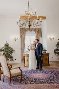Ein Paar steht in einem eleganten, mit Pflanzen dekorierten Raum, der perfekt für eine Hochzeit geeignet ist, vor einem von Vorhängen umrahmten Fenster. Die Frau trägt ein weißes Kleid und der Mann einen dunklen Anzug. Darüber hängt ein Kronleuchter und ein gemusterter Teppich bedeckt den Boden.