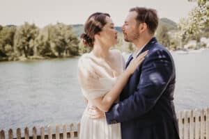 Ein Paar umarmt sich am Ufer eines Sees und versinkt in seinem Hochzeitsglück. Die Frau im weißen Kleid legt liebevoll ihre Hände auf die Schultern des Mannes. Der Mann, in einen marineblauen Anzug gekleidet, blickt sie hingebungsvoll an. Bäume und Hügel rahmen ihren Moment unter dem klaren Himmel ein.