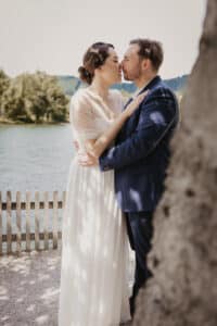 Ein Paar in formeller Kleidung umarmt und küsst sich an einem Seeufer und feiert seine Hochzeit. Die Frau trägt ein weißes Kleid und der Mann einen dunklen Anzug. Sie stehen neben einem Holzzaun mit Bäumen und Wasser im Hintergrund, aufgenommen im sanften Tageslicht.