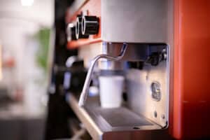 Nahaufnahme einer Kaffeemaschine mit rotem und silbernem Gehäuse, perfekt für die Hochzeitsplanungstage. Unter dem Ausguss wartet ein weißer Pappbecher darauf, gefüllt zu werden. Verschiedene Knöpfe und Regler weisen auf die Vielseitigkeit hin. Der unscharfe Hintergrund schafft eine gemütliche Innenatmosphäre, die sich ideal für die Hochzeitsvorbereitungen eignet.