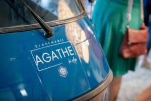 Nahaufnahme eines blauen Fahrzeugs mit dem Aufdruck „Eventmobil AGATHE by dfp“, perfekt für Hochzeitsfeiern. Ein Logo, das an ein Spielkartensymbol erinnert, ist sichtbar. In der Nähe steht eine Person in einem grünen Kleid, die möglicherweise an der Hochzeitsplanung beteiligt ist, und ist teilweise sichtbar.