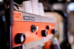 Nahaufnahme einer orangefarbenen Rancilio-Espressomaschine mit Bedienknöpfen und Schaltern. Ideal für eine Café-Umgebung, perfekt für ein Hochzeitsplanungstreffen, wobei der sanft fokussierte Hintergrund auf intime Feiern und Diskussionen zum Thema Heiraten hindeutet.