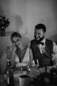 Ein glückliches Paar sitzt eng an einem wunderschön dekorierten Tisch, der Mann lächelt und umarmt die Frau zärtlich. Sie gestikuliert verspielt in Richtung ihres Gesichts und fängt einen warmen Moment ein, der den Zauber der Liebe und des Heiratens ausstrahlt, wobei Blumen und elegantes Geschirr den Vordergrund schmücken.