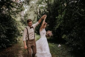 Ein Paar tanzt fröhlich einen Waldweg entlang, umarmt vom Geist des Heiratens. Der Bräutigam trägt eine braune Weste und Fliege, während die Braut ein weißes Kleid trägt und einen Blumenstrauß hält. Bäume und Grün umgeben sie und schaffen eine romantische, natürliche Kulisse, die ideal für ihre Hochzeit ist.