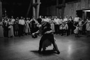 Ein Paar tanzt in der Mitte einer Scheune und fängt einen Hochzeitsmoment ein, während die Menge zusieht. Der Mann lässt die Frau tief sinken, umgeben von Holzbalken. Die verkleideten Erwachsenen und ein Kind schaffen eine festliche Atmosphäre. Dieses Schwarz-Weiß-Bild verkörpert wunderschön die Magie des Heiratens.