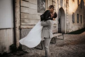 Ein Mann in einem hellgrauen Anzug hebt eine Frau in einem fließenden weißen Kleid und einer schwarzen Jacke vor einem alten Steingebäude hoch. Momente purer Freude während ihrer Hochzeit. Sie umarmen sich freudig auf einem Kopfsteinpflasterweg neben einer Holzbank und fangen die Essenz von Liebe und Feier ein.