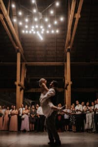 Ein Paar tanzt unter einem Kronleuchter in einem rustikalen Hochzeitslokal, wobei der Bräutigam die Braut hochhebt. Gäste umringen sie und schauen glücklich zu. Die Atmosphäre ist warm und festlich und spiegelt die Freude des Heiratens in dieser zauberhaften Umgebung wider.
