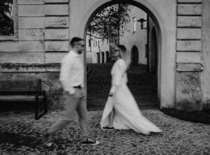 Ein Schwarzweißfoto zeigt einen Mann und eine Frau, die auf einem Kopfsteinpflasterweg unter einem Torbogen aneinander vorbeigehen, möglicherweise auf dem Weg zu ihrer Hochzeit. Die Frau trägt ein fließendes Kleid und Turnschuhe, während der Mann Hemd und Hose trägt. Sie sind leicht verschwommen, was Bewegung suggeriert.
