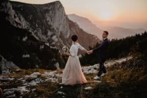 Ein Paar im Hochzeitskleid steht bei Sonnenuntergang auf einem Bergpfad und hält einen Moment fest, den es für immer in Erinnerung behalten wird. Die Braut hält einen Blumenstrauß und die Hand des Bräutigams, während sie zum Horizont blicken. Dramatische Klippen und eine strahlende Sonne bilden die Bühne für ihre Romanze – ein exquisites Hochzeitsziel.